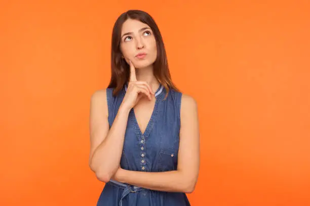 Photo of Pensive brunette woman in denim dress looking up with thoughtful doubtful expression