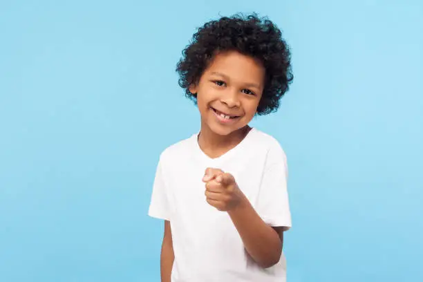 Photo of Hey you! Portrait of cheerful funny little boy with curly hair pointing finger to camera and smiling, child making choice indicating at you