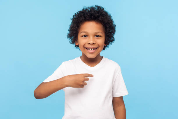 This is me. Portrait of happy preschool curly boy in T-shirt joyfully looking at camera and pointing to himself This is me. Portrait of happy preschool curly boy in T-shirt joyfully looking at camera and pointing to himself, proud of own success, egoistic child. indoor studio shot isolated on blue background achievement aiming aspirations attitude stock pictures, royalty-free photos & images