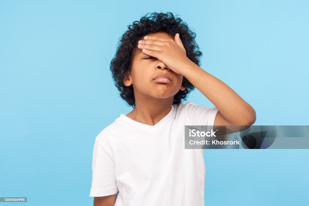 Facepalm. Portrait of forgetful upset little boy with curls covering face with hand and expressing sorrow Facepalm. Portrait of forgetful upset little boy with curls covering face with hand and expressing sorrow regret for mistake, forgot to do homework. indoor studio shot isolated on blue background Child Stock Photo