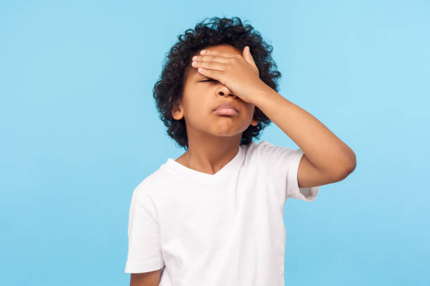 Facepalm. Portrait of forgetful upset little boy with curls covering face with hand and expressing sorrow Facepalm. Portrait of forgetful upset little boy with curls covering face with hand and expressing sorrow regret for mistake, forgot to do homework. indoor studio shot isolated on blue background embarrassment stock pictures, royalty-free photos & images