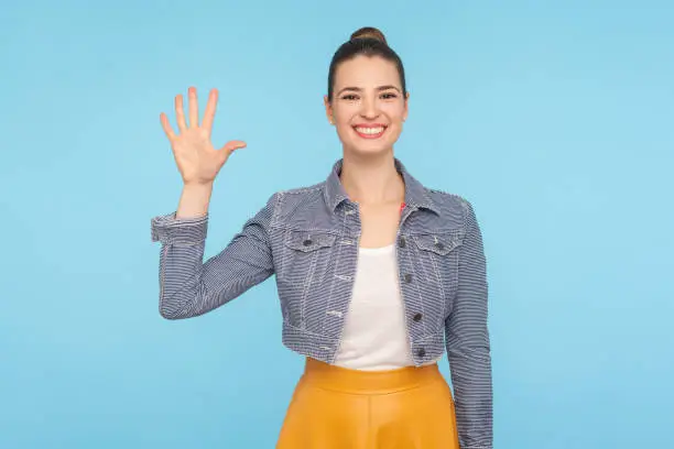 Hello! Portrait of friendly sociable woman in stylish casual clothes gesturing hi, welcoming with raised hand and looking at camera with affable smile. indoor studio shot isolated on blue background