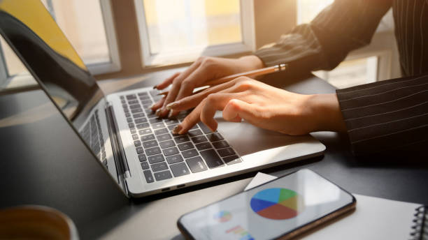 istock Side view of female working on laptop while looking business chart on smartphone 1204522577
