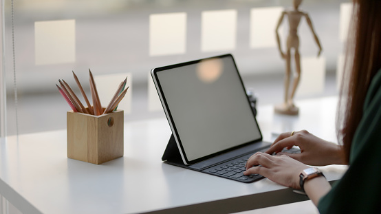 Cropped shot of female designer typing on blank screen tablet with wooden figure and coloured pencils on white table in simple co working space