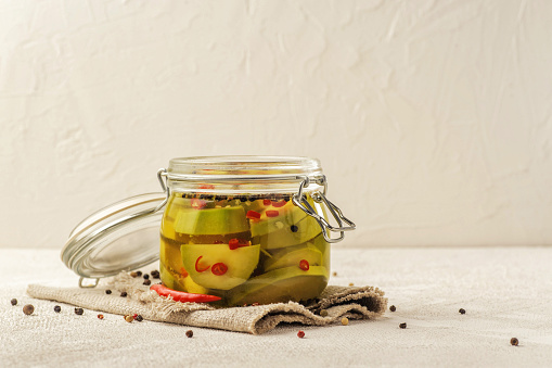 Fermented avocado vegetables with red chili peppers. Glass jar on a light background. Natural linen fabric.