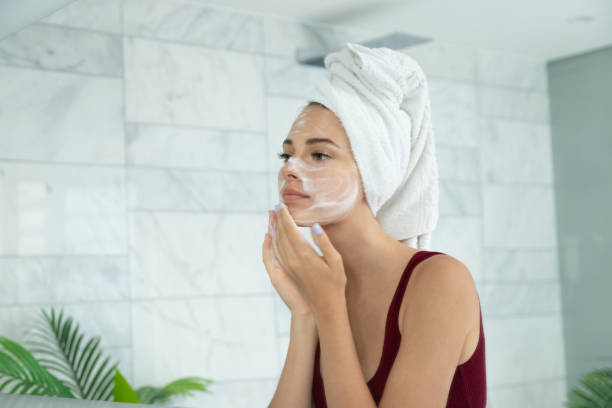 Beautiful woman applying moisturizing skin lotion Beautiful  woman applying facial  foam  standing front of mirror in bathroom and cleansing, moisturising and protection skin. cheek photos stock pictures, royalty-free photos & images