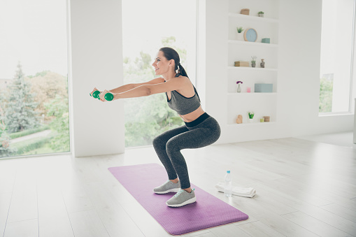 Full size profile side photo of joyful energetic sportswoman want be strong girl do aerobics sporty exercise make squats with her hands, holding green dumbbells stand on purple mat in house like gym
