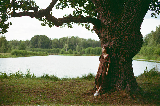 Young Caucasian woman standing the old oak by the lake