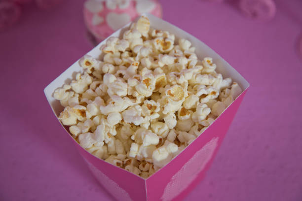 A wide classic box of theater popcorn isolated on pink . Popcorn spilled from a striped box . Three vintage-style boxes of popcorn in a carnival themed still life with arcade game pink tickets palomitas de maíz derramadas de una caja a rayas. tres cajas de estilo vintage de palomitas de maíz en un bodegones temático de carnaval con entradas rosas juego de arcade. una amplia caja clásica de palomitas de maíz de teatro aisladas en rosa.