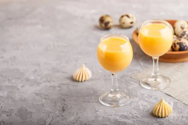 Sweet egg liqueur in glass with quail eggs and meringues on a gray concrete background. Side view, copy space.