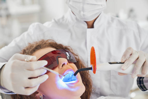 Attractive lady doing whitening procedure in dental clinic Attractive lady with curly hair lying in dental chair in protective glasses with opened mouth while while male doctor in white glasses doing whitening procedure. Concept of dental care dental light stock pictures, royalty-free photos & images