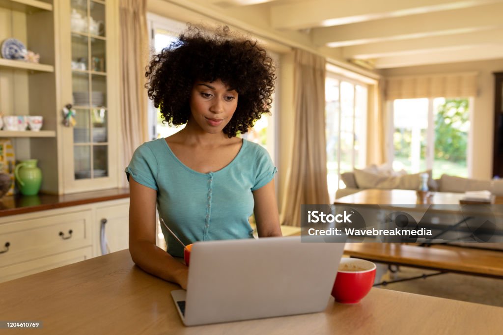Young woman using computer in the kitchen Weekend at home together. Front view of a mixed race woman sitting in the kitchen having breakfast and using her computer laptop In Front Of Stock Photo