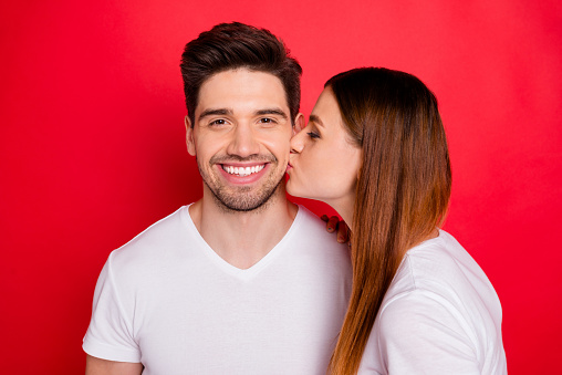 Photo of redhair positive cute nice pretty couple of married people with wife in white t-shirt kissing her husband cheek smiling toothily, showing their tender love isolated bright red color background