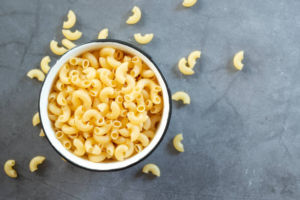 raw elbow macaroni in enamel bowl raw elbow macaroni in enamel bowl on dark cement table background. Macaroni stock pictures, royalty-free photos & images