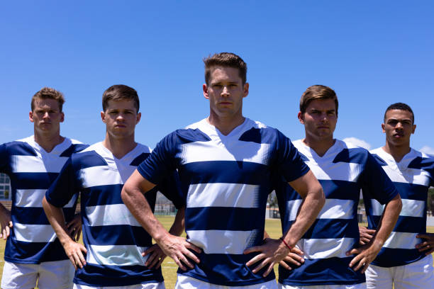 Rugbymen team standing Front view of a multi-ethnic group of male rugby players wearing a team strip, standing on a pitch with hands on hips, looking straight to camera before playing a match, with blue sky in the background rugby team stock pictures, royalty-free photos & images