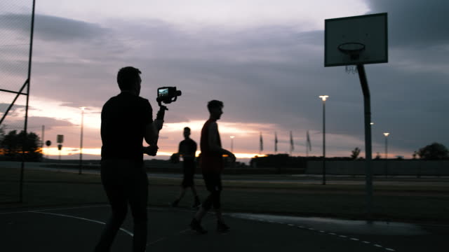 MS Man filming basketball players shooting hoops on outdoor basketball court at dusk