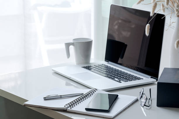 istock Mockup smartphone on open notebook with laptop, glasses,box and cup on white wooden table. 1204621020