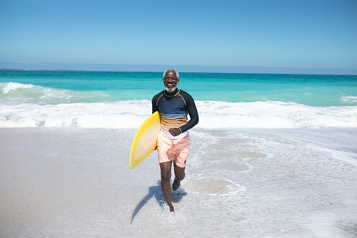 Front view of a senior African American man on a beach in the sun, carrying a surfboard under his arm, running and looking to camera, smiling, with blue sky and sea in the background