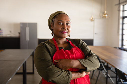 Portrait of a confident African American woman wearing a red apron at a cookery class, standing in a restauarant with her arms crossed, looking to camera and smiling