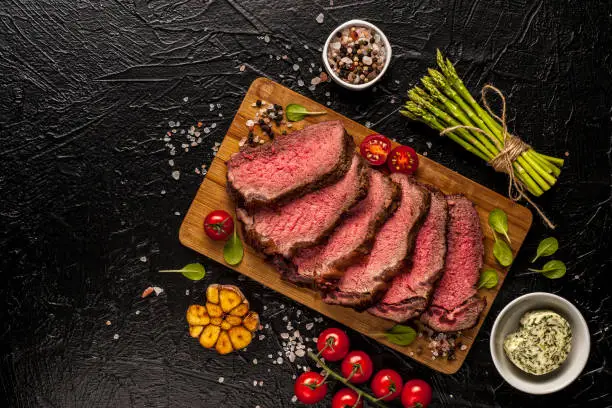 Roast beef. A large piece of meat with fresh vegetables is cut on a board and ready to eat. Black background. Top view. Close up and horizontal orientation. "n