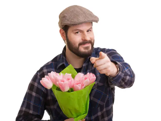 Bearded young handsome man with pointed indexfinger holding a bouquet of pink tulips, isolated on white background. Mothers day, Valentines day, Easter and surprise concept.