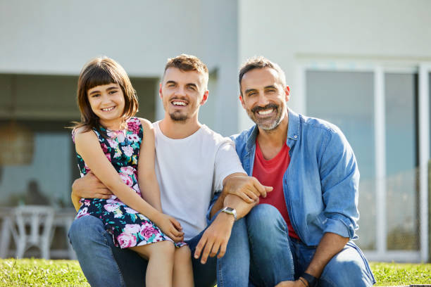Portrait of homosexual men with daughter at yard Portrait of homosexual men with daughter at back yard. Smiling family is sitting against house. They are outdoors on sunny day. gay daddy stock pictures, royalty-free photos & images