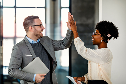 Coworkers celebrating achievement at office. Celebrating success. Happy young man standing in office and giving high five to his colleagues. Two business people high-five. Job well done