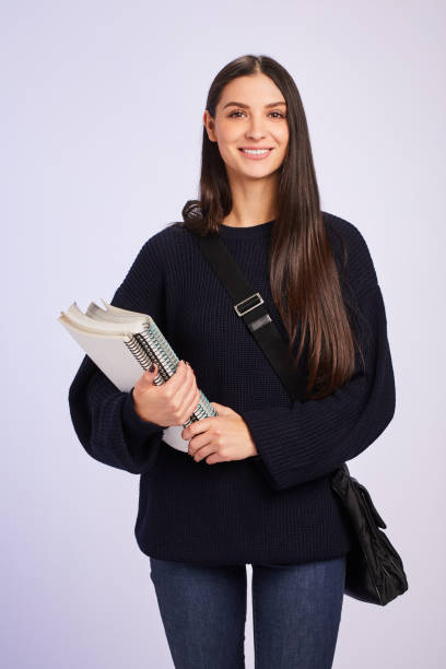 Studio portrait of a young female university student smiling while carrying a bag and textbooks against a colored background