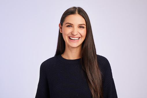 Studio portrait of a young woman with long brown hair and wearing a sweater laughing while standing against a colored background