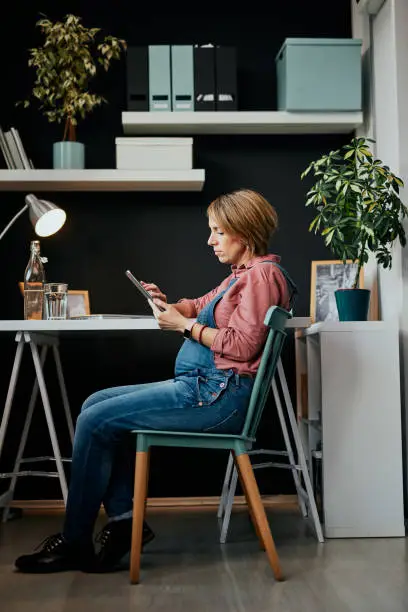 Side view of cute smiling caucasian pregnant woman sitting in home office and using tablet.