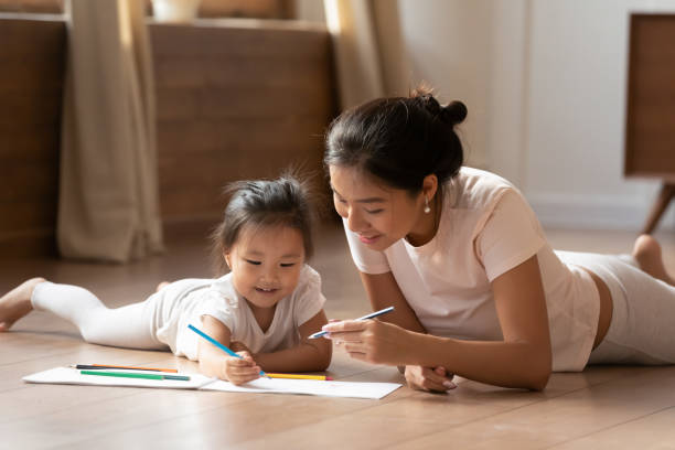 Young Asian mother and little daughter drawing together Smiling Vietnamese young mom and little biracial daughter lie on warm wooden heated floor in living room drawing painting together, happy ethnic mother or nanny relax play with small Asian girl child asian nanny stock pictures, royalty-free photos & images
