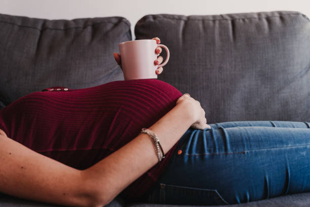 porträt der jungen schwangeren frau zu hause liegend auf dem sofa und hält eine tasse auf dem bauch - caffeine free stock-fotos und bilder