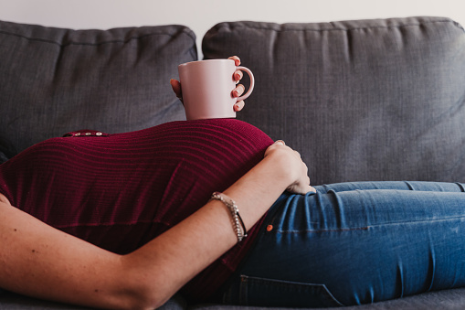 retrato de joven embarazada en casa acostado en el sofá y sosteniendo una taza en el vientre photo