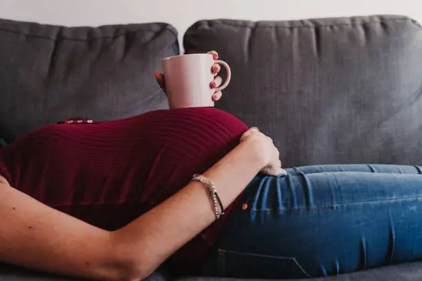 Photo of portrait of young pregnant woman at home lying on the sofa and holding a cup on belly