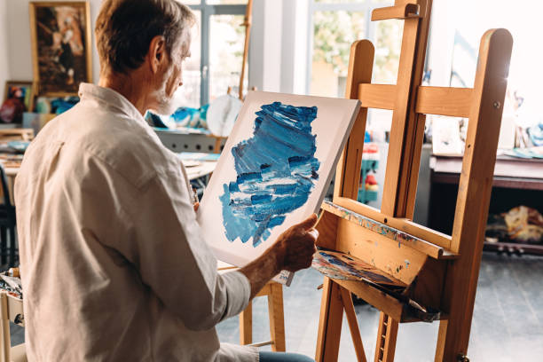 istock Back view of male painter holding a canvas with abstract picture sitting at easel 1204687286