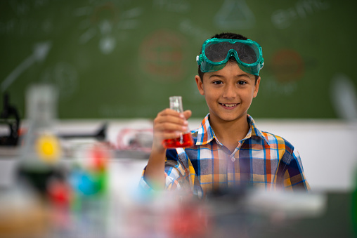 A male elementary student sits at his desk in class holding up a beaker with an orange chemical/liquid inside it. He is smiling and has protective goggles on his forehead. The desk in front of him has molecule models on it and there is a blackboard in the background with writing on it.