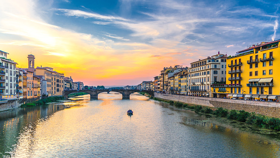 St Trinity Bridge stone bridge and boat on Arno River water and embankment promenade with buildings in historical centre of Florence city, bright blue orange evening sky clouds, Tuscany, Italy