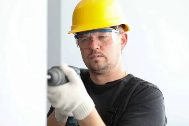 Close-up view of concentrated male drilling hole in wall. Professional builder wearing yellow protective helmet and eyewear. Construction and foreman concept