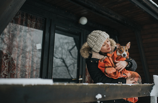 young woman enjoying winter holiday at village with her dog. They are standing iin front of wooden house enjoying the view.Winter time.