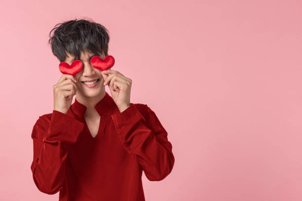 Young man in red shirt holding red heart in front of eye with pinky background stock photo