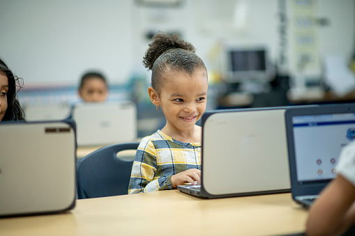 A group of multi-ethnic elementary students sit at desk in a computer lab, with a laptop in front of each of them.  They are each dressed casually and smiling.  The focus is on a sweet girl of African decent.