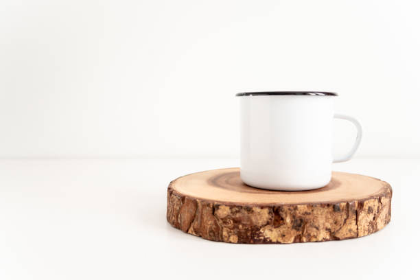 Enamel mug mock up with wooden cut tree section on a white table. Boho design of tin cup on white background with copy space for image or text
