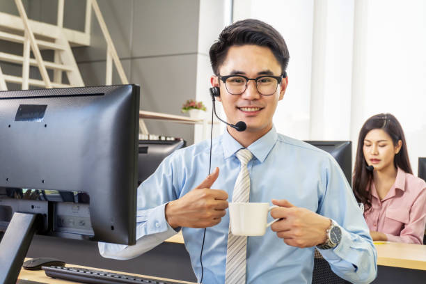 centre d’appel, portrait de l’opérateur de téléphone masculin heureux de soutien à la clientèle de sourire au lieu de travail, l’homme gai de centre d’appel travaillant du bureau parlant avec le client et buvant le café - women on the phone headset service photos et images de collection