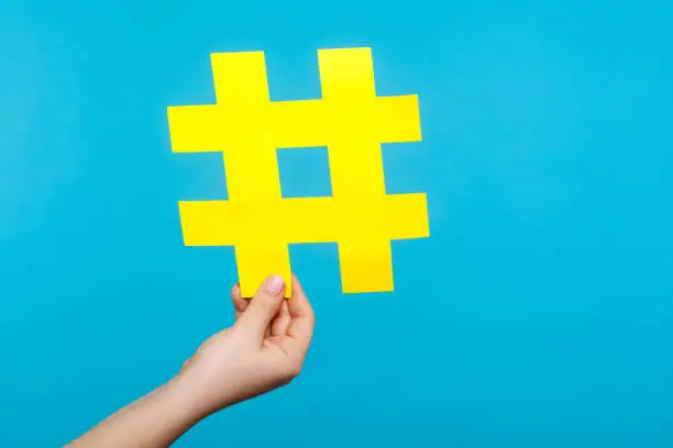 Photo of Closeup of female hand holding big yellow paper hashtag sign, hash symbol of internet popularity. isolated on blue background