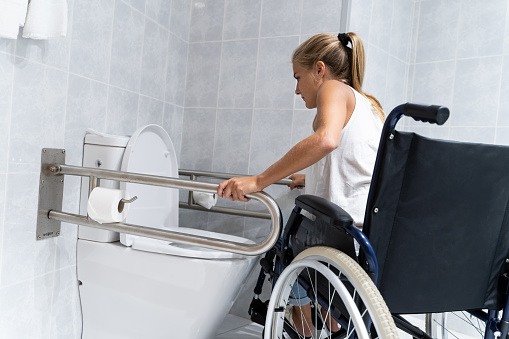 Blonde girl holding on to rails at the toilet trying to get up with her arms from a wheelchair to go to the bathroom