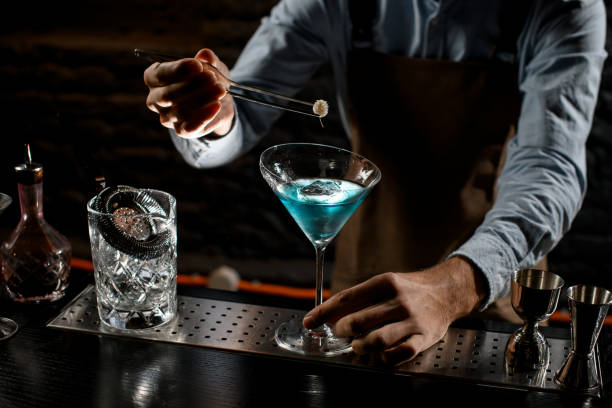 istock Professional bartender decorating a blue alcoholic cocktail in a martini glass with a spikelet by tweezers 1204783332