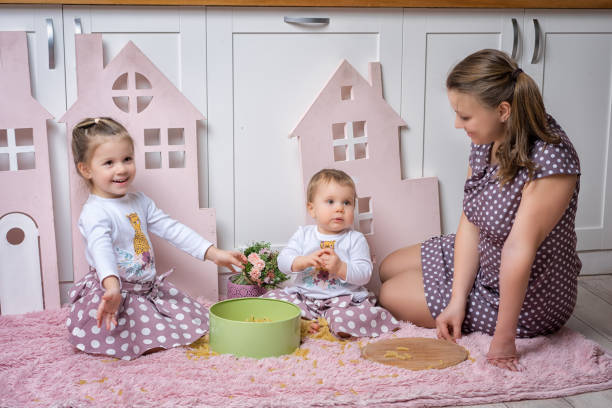 madre y hermanas niñas de 1 y 3 años de edad se sentan en el suelo en la cocina y juegan con pasta seca. - 2 3 years children only group of people enjoyment fotografías e imágenes de stock