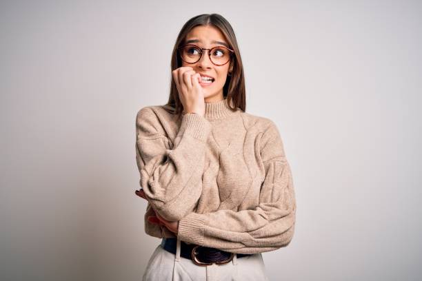 カジュアルなセーターと眼鏡をかけている若い美しいブルネットのビジネスウーマンは、口を噛む爪に手を置いてストレスと緊張を見て立っています。不安の問題. - shy ストックフォトと画像