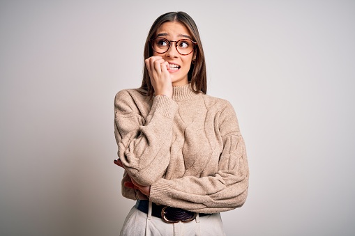 Young beautiful brunette businesswoman wearing casual sweater and glasses standing looking stressed and nervous with hands on mouth biting nails. Anxiety problem.