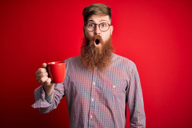 istock Redhead Irish man with beard drinking a cup of hot coffee over red background scared in shock with a surprise face, afraid and excited with fear expression 1204794431